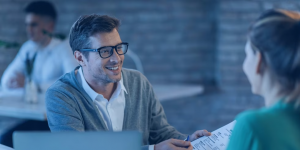 Capa de artigo: Investidor moderado. Homem sentado à mesa enquanto conversa com sua assessora de investimentos