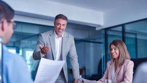 Capa de Artigo: Assessoria de Investimentos. Descrição: Homem branco em pé ao lado de uma mesa, conversando com homem e mulher sobre negócios, em uma sala de reunião